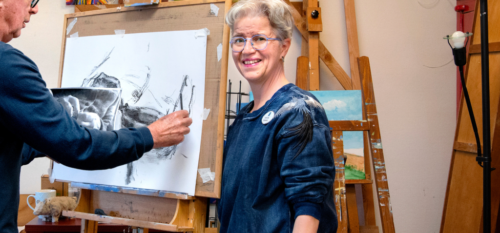 Logopedist Laura Bas staat naast een deelnemer van het afasie atelier. Deze deelnemer schetst met houtskool op een doel dat op een schildersezel staat.