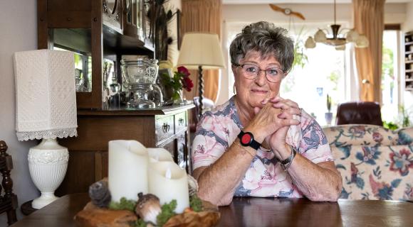 Mevrouw zit aan de keukentafel in haar woning en heeft een armband om met een alarmknop voor personenalarmering.