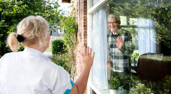Medewerker van de thuiszorg zwaait naar een meneer die thuis in zich woonkamer staat en haar ziet vanuit zijn raam.