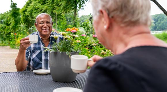 Vrijwilliger drinkt een kop koffie met een bewoner van een woonzorglocatie, buiten in de tuin.
