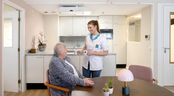 Bewoner van woonlocatie Betsy Perk zit aan tafel in een appartement terwijl een zorgmedewerker koffie serveert. Zorg en ondersteuning zijn dichtbij, terwijl bewoners zelfstandig wonen.
