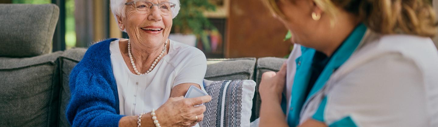 Omring verpleegkundige thuis op de bank bij een mevrouw die een glucosesensor op haar arm heeft.