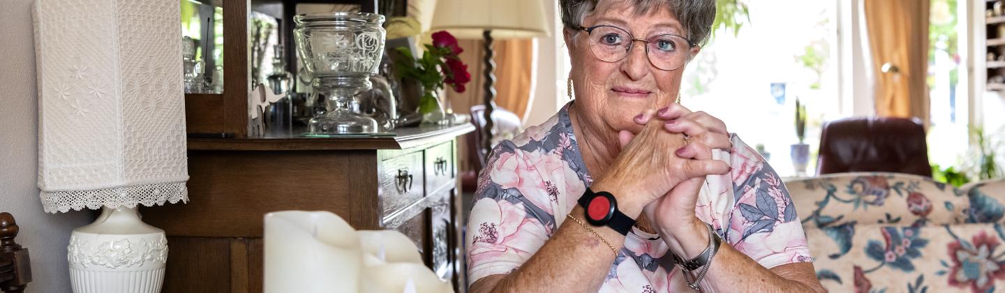 Mevrouw zit aan de keukentafel in haar woning en heeft een armband om met een alarmknop voor personenalarmering.