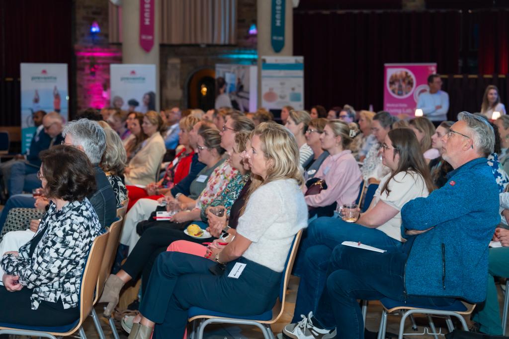 Overzicht van een volle zaal toeschouwers die geïnteresseerd luisteren naar de persoon (buiten beeld) op het podium.