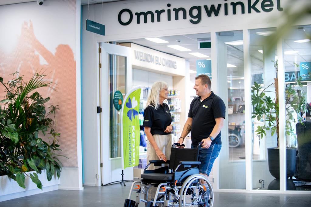 Meneer en mevrouw staan met een rolstoel voor de Omringwinkel