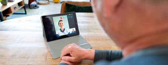 Over de schouder van een meneer meekijkend naar een iPad op tafel, waarop de verpleegkundige zichtbaar is.