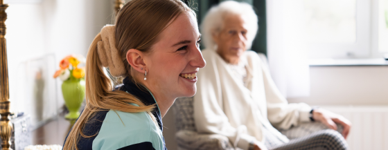 Een medewerker van Omring is in gesprek en lacht naar iemand buiten beeld. Op de achtergrond zit een bewoner in haar stoel.