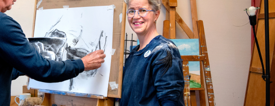 Logopedist Laura Bas staat naast een deelnemer van het afasie atelier. Deze deelnemer schetst met houtskool op een doel dat op een schildersezel staat.