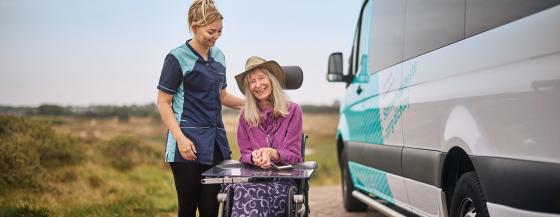 Medewerker van Omring en cliënt in een rolstol staan naast de Omringbus in de duinen van Den Helder.